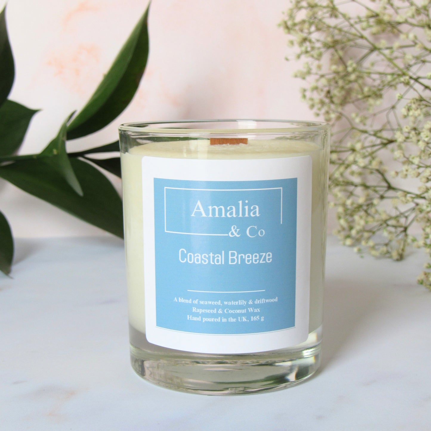 Coastal Breeze wood wick candle with sea salt and driftwood scent in a clear jar.A white candle with a blue label that reads 'Amalia & Co Coastal Breeze', set in a clear glass jar. The candle is accompanied by green foliage and white flowers in the background.