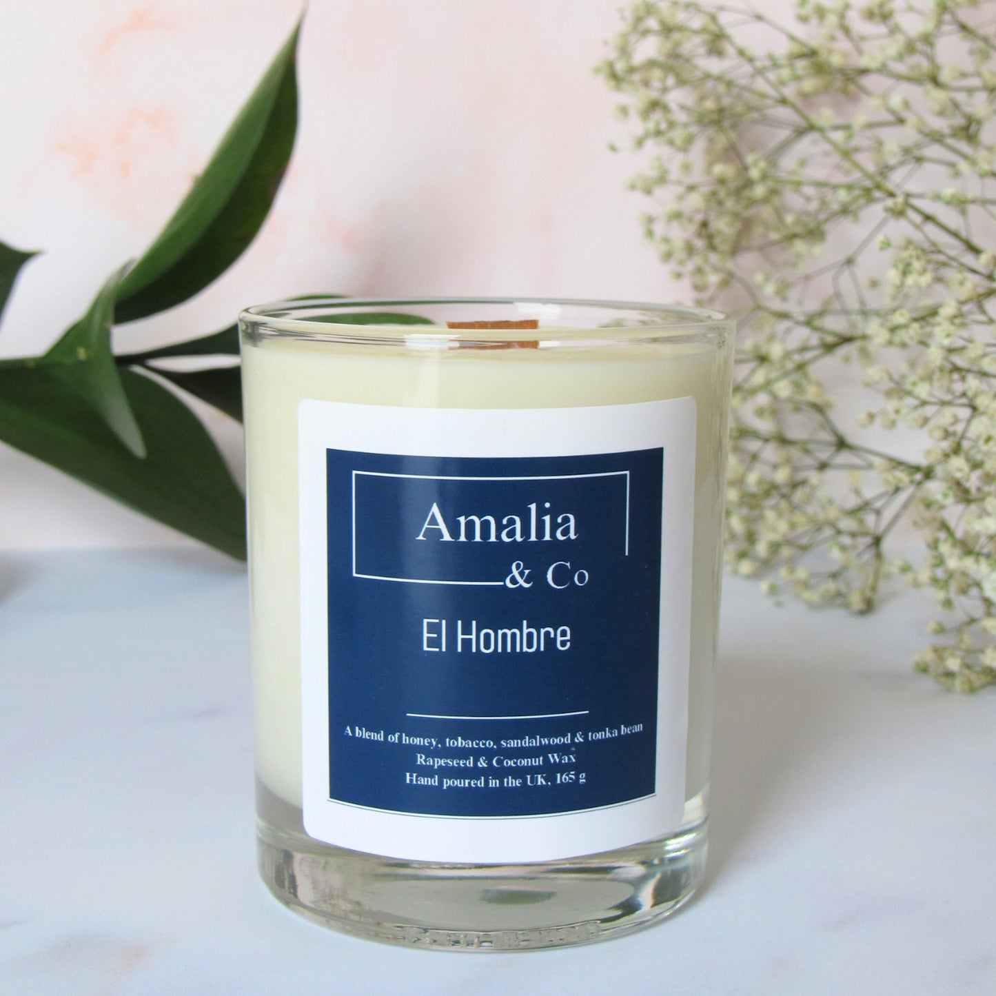 Sophistication in a jar. El Hombre features bold tobacco, honey, and sandalwood. The ultimate luxury masculine scent. Discover our wood wick collection.A white candle with a blue label that reads 'Amalia & Co - El Hombre'. The candle is placed on a white surface with green leaves and white flowers in the background.