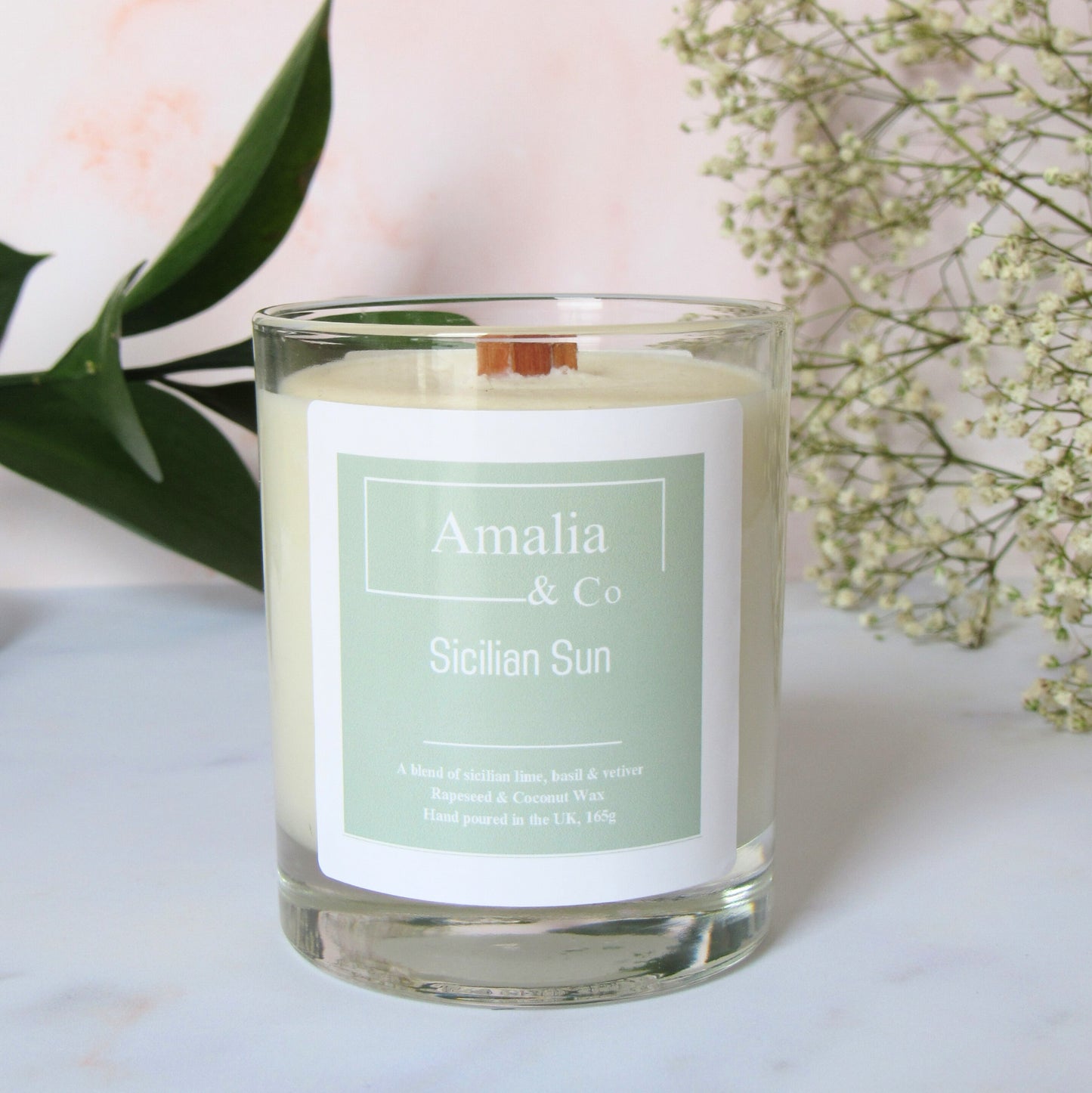 A white candle labeled 'Amalia & Co Sicilian Sun' with a wooden wick, housed in a clear glass jar. The label has green accents and the candle is placed on a white surface with green leaves and white flowers in the background.